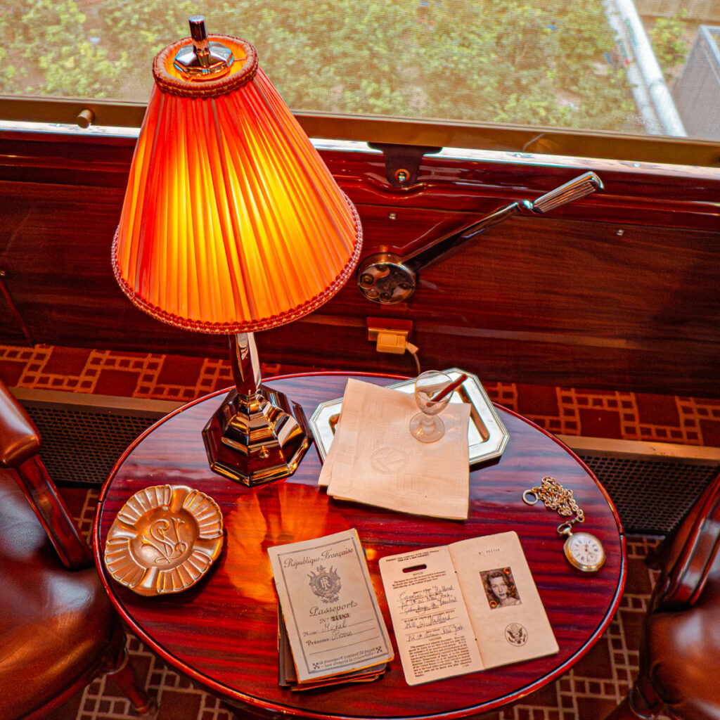 Vue d'une table ronde richement décorée avec des accessoires tels qu'un passeport d'espionne et d'autres objets, disposée dans une voiture de l'Orient Express lors de l'exposition 'Il était une fois l'Orient Express' à l'Institut du Monde Arabe à Paris en 2014, avec une lampe chic, créant une ambiance intrigante et mystérieuse, comme si les voyageurs venaient juste de quitter la pièce.