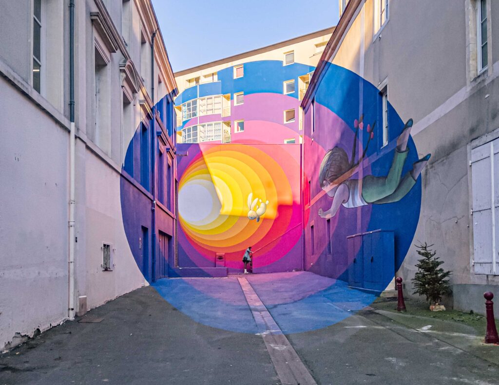 Cette image capture le street art intitulé "Rabbit Hole", une œuvre magistrale du célèbre artiste de rue Seth. La fresque est une anamorphose qui dépeint une petite fille dans un trompe-l'œil coloré, créant une brèche vers un univers imaginaire. L'image déformée n'acquiert toute sa signification que lorsqu'elle est vue d'un point précis, aisément repérable par le pavé manquant à l'entrée de l'impasse Jankowski. 

Cette perfection visuelle est un émerveillement pour les yeux, offrant un effet de perspective saisissant et empli de poésie. On y voit une petite fille suivant un lapin blanc vers un tunnel arc-en-ciel, peut-être une allusion à Alice au Pays des Merveilles. L'invitation est irrésistible : on se sent appelé à suivre l'enfant et à découvrir ce qui se cache derrière ce terrier enchanté.