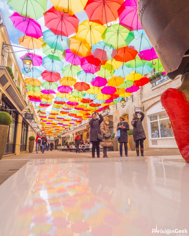 Des passants prennent en photo les parapluies multicolores du Village Royal à Paris
