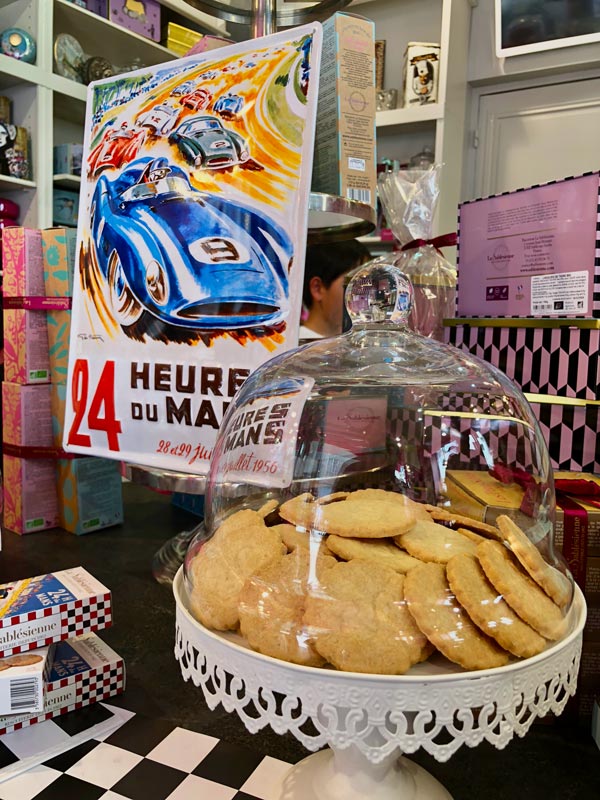 Une photo de l’intérieur de la boutique La Sablésienne au Mans, spécialisée dans les biscuits sablés. On voit une boîte en métal décorées avec le logo et l’affiche des 24 Heures du Mans, contenant des biscuits sablés de la marque. On voit aussi des biscuits sablés sous verre. L’ambiance est chaleureuse et gourmande.