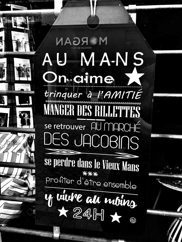 Une photo d’un panneau en bois avec l’inscription “Au Mans on aime” en lettres blanches. Le panneau est accroché derrière une vitre dans une boutique du centre ville du Mans. 