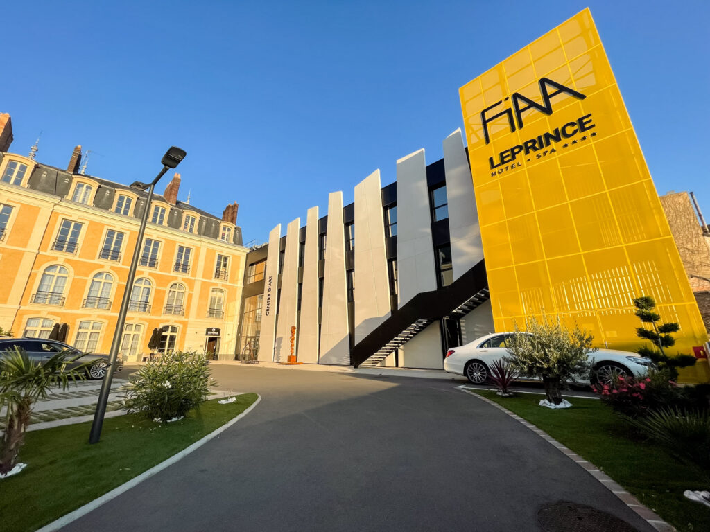 Cette image, prise par ParisianGeek, présente l'Hôtel Le Prince et le bâtiment du FIAA au Mans. L'architecture élégante de l'Hôtel Le Prince, avec son design classique et ses détails raffinés, contraste avec le bâtiment moderne du FIAA, illustrant la diversité architecturale de la ville. La scène est capturée avec une précision qui met en valeur la beauté et l'unicité de chaque bâtiment, tout en montrant comment ils coexistent harmonieusement dans l'environnement urbain. C'est une image qui célèbre le mélange dynamique du vieux et du nouveau qui fait partie intégrante de l'identité du Mans.