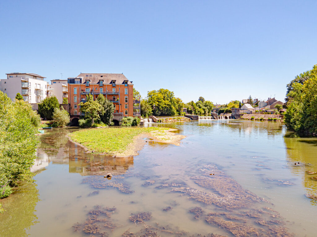 Une photo d’une rivière appelée l’Huisne, qui traverse la ville du Mans. La photo montre un endroit nommé le Gué de Maulny, où la rivière forme un plan d’eau calme et verdoyant. On y voit des arbres, des plantes aquatiques et des reflets du ciel. Au loin, on aperçoit un pont en pierre qui enjambe la rivière. La photo est prise par ParisianGeek