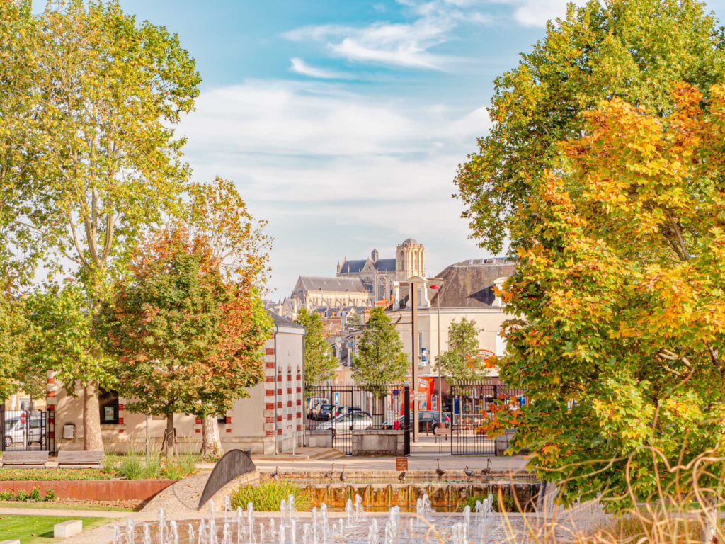 Une photo de la cathédrale du Mans vue depuis le parc Théodore Monod. On voit la façade et la tour sud de la cathédrale, qui se détachent sur le ciel bleu. Le parc est verdoyant et roux, avec des bancs et des lampadaires. On voit aussi un jet d’eau au premier plan.