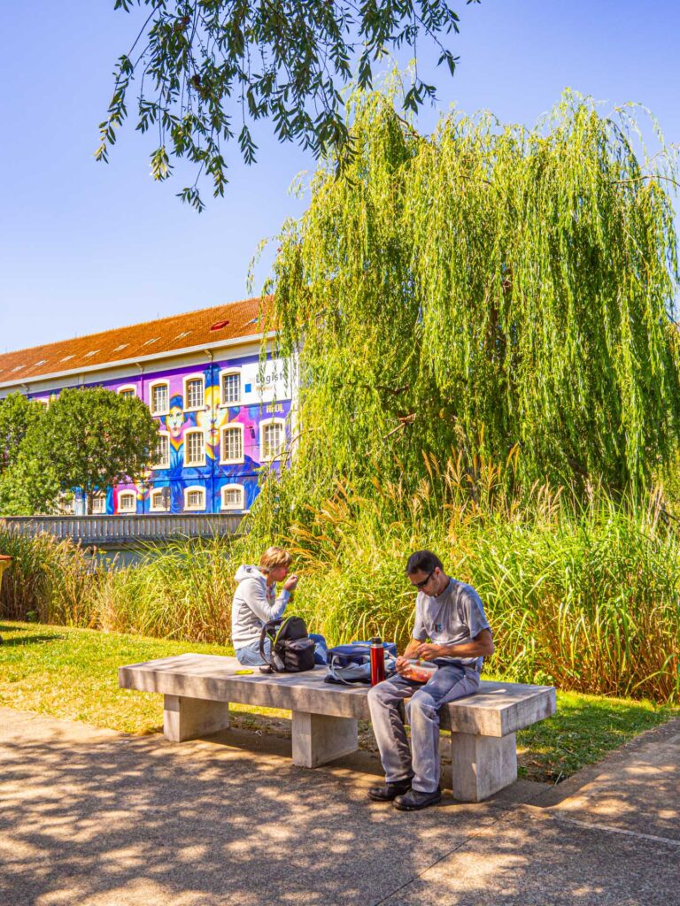 Une photo capturant une scène paisible à l'Île aux Planches, Le Mans, France, prise par ParisianGeek. L'image dépeint un groupe de personnes profitant d'une journée ensoleillée dans le parc, certains assis sur des bancs à l'ombre des arbres, d'autres se promenant le long des allées sinueuses. Les couleurs vives de la nature - le vert des arbres et l'herbe, le bleu du ciel - créent une atmosphère sereine et rafraîchissante. Au loin, on peut apercevoir les contours de la ville, avec des bâtiments pittoresques qui ajoutent une dimension historique à la scène. L'image capture parfaitement le charme tranquille de l'Île aux Planches, un lieu de détente et de loisirs au cœur de la ville animée du Mans.