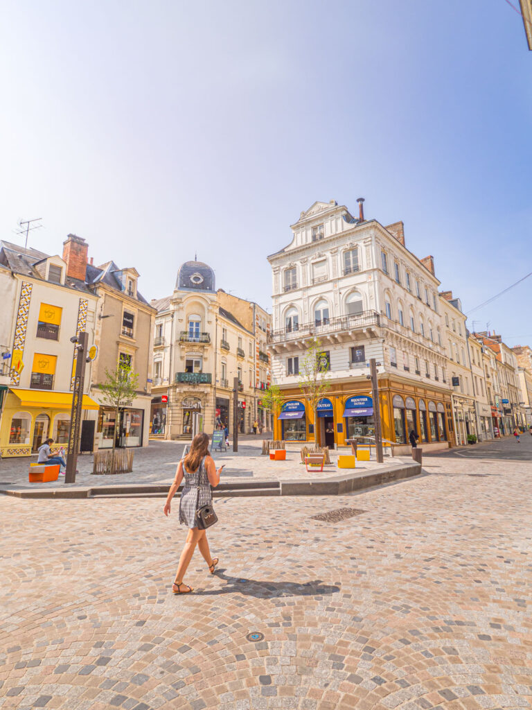 Une photo d’une place publique nommée Place Saint-Nicolas, située dans le centre historique du Mans. La place a été rénovée récemment et présente un aspect moderne et lumineux. On y voit des bancs, des lampadaires, des arbres et des fleurs. Au fond de la photo, on aperçoit l’église Saint-Nicolas, un monument historique du XIIe siècle. Au premier plan, une femme marche sur le pavé