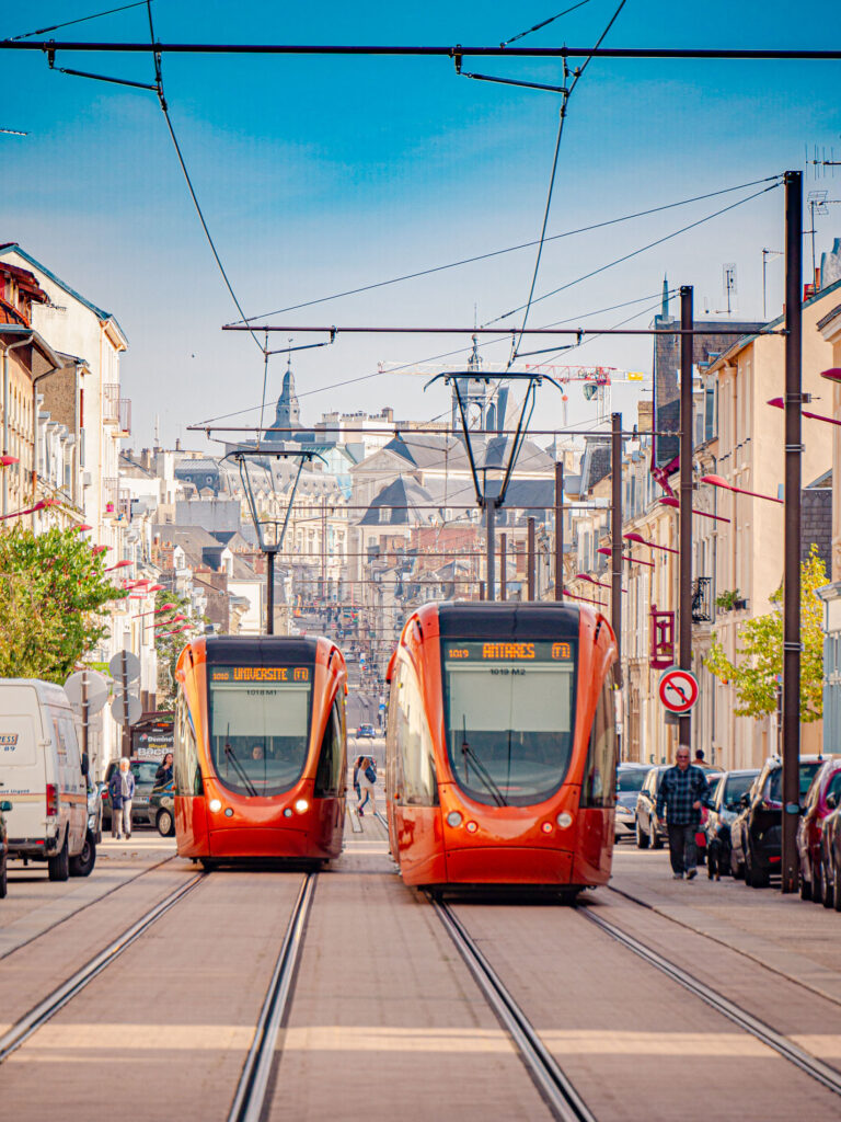 Une photo de deux tramways du Mans circulant Rue Gambetta, l’un en direction d’Université et l’autre en direction d’Antarès. Les tramways sont de couleur orange.