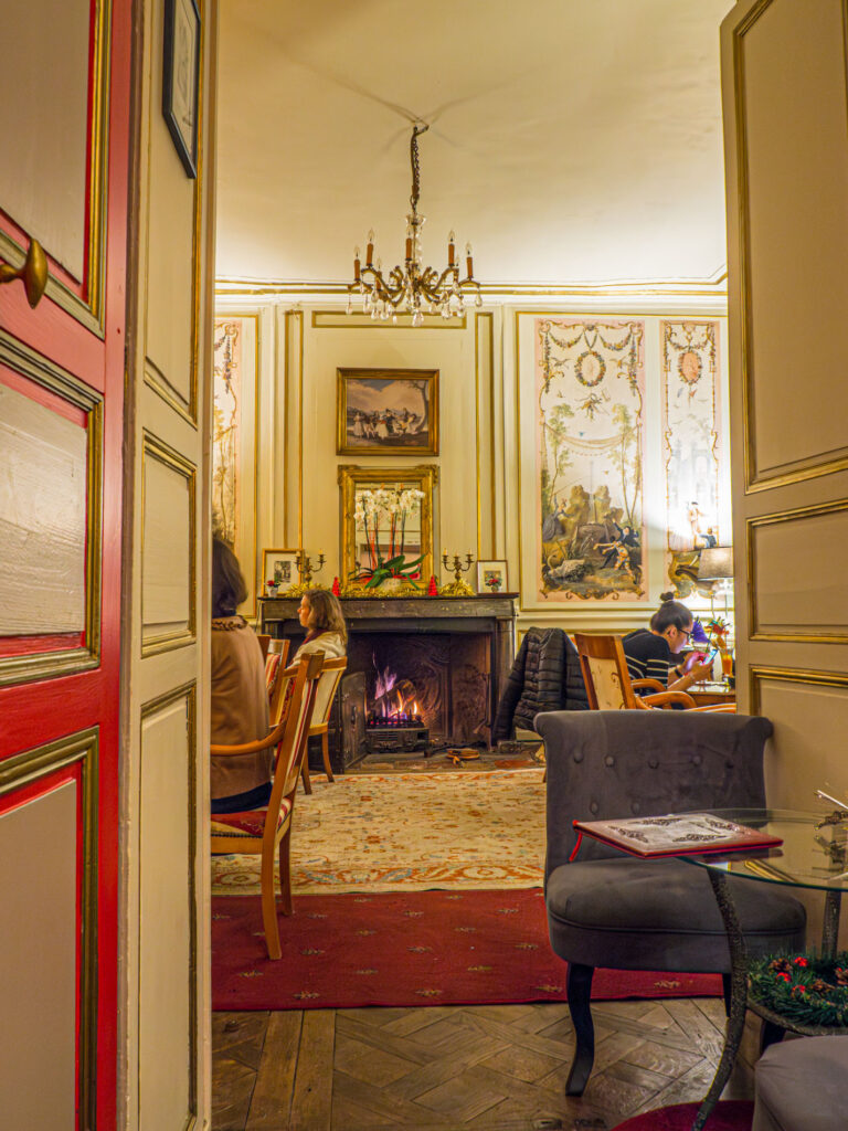 Une photo de l’intérieur du bar à cocktails Le Saint-Flaceau au Mans, dans un style 18e. On voit le comptoir en bois orné de sculptures et de dorures, ainsi que des bouteilles et des verres anciens. Les murs sont recouverts de tapisseries et de tableaux. Des lustres en cristal et des bougies éclairent la pièce. Des fauteuils et des canapés en velours rouge invitent à la détente. Message reçu. Un texte alternatif avec texte possible est : Une photo de l'intérieur du bar à cocktails Le Saint-Flaceau au Mans, dans un style 18e. On voit le comptoir en bois orné de sculptures et de dorures, ainsi que des bouteilles et des verres anciens. Les murs sont recouverts de tapisseries et de tableaux. Des lustres en cristal et des bougies éclairent la pièce. Des fauteuils et des canapés invitent à la détente.